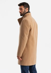 Beige Wollmantel mit hohem Kragen, taillierter Passform und glatter Textur, kombiniert mit dunklen Jeans, von hinten betrachtet.