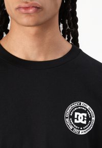 Juodas medvilninis marškinėliai su apvaliu baltu logotipu, kuriame yra tekstas "DC Shoe Company" ir įkūrimo metai. Apvalus kaklas ir trumpomis rankovėmis.