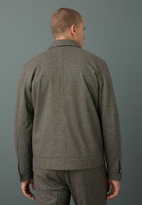 Chaqueta gris a cuadros con cuello, que presenta puños con botones y un corte recto. El material parece texturizado con un patrón sutil.