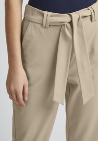 Beige Hosen mit glatter Textur, die einen breiten Gürtel haben, der zu einer Schleife gebunden wird, sowie genähte Gürtelschlaufen in der Taille.