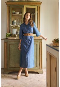 Žena v modrých midi šatech na knoflíky s hnědým pásem a balerínkami stojí v rustikální kuchyni vedle zelené dřevěné skříně.