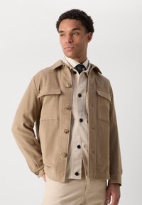 Veste beige à boutons avec deux poches avant, col classique et boutons marron, superposée sur une chemise claire et une cravate. Texture de tissu lisse.