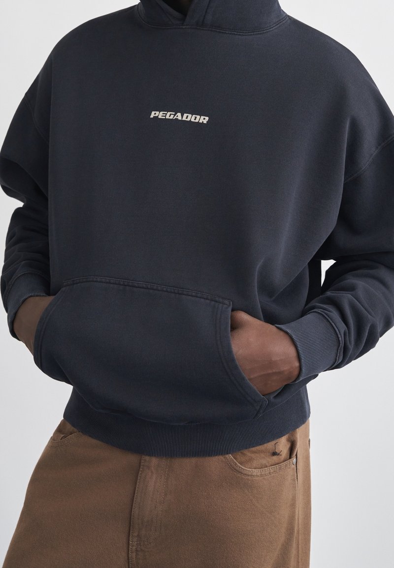 Sweat à capuche bleu marine foncé avec petit texte "PEGADOR" sur la poitrine, poche kangourou à l'avant, porté par une personne avec les mains dans la poche.
