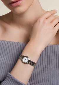 Gold- und silberfarbene achteckige Uhr mit weißem Ziffernblatt, Metallarmband, getragen am Handgelenk in der Nähe eines gerippten hellgrauen Oberteils.