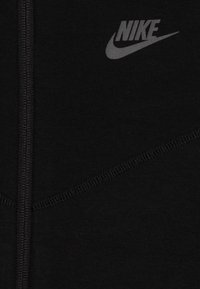 Svart zip-up-jacka med en slät textur, som har en grå Nike-logotyp på den övre vänstra sidan och en subtil sömdetalj över tyget.