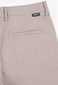 Jasno beżowe spodnie z tylnią kieszenią, wykonane z gładkiego materiału. Posiadają czarną naszywkę PROSTO przyszytą w pasie.