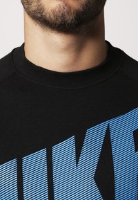 Černá mikina se modro pruhovaným logem. Materiál je žebrovaný, má kulatý výstřih a viditelné švy na okrajích.