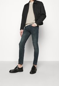 Svart bomberjacka, beige tröja, mörkblå slim-fit jeans och svarta läderskor med spännen. Enkelt design, inga synliga mönster.