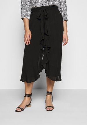 Kobieta w czarnej spódnicy midi z przednią falbaną i wiązaniem w kokardę, zestawiona z czarnymi sandałami z zawiązywaniem wokół kostki oraz wzorzystą bluzką z długim rękawem.