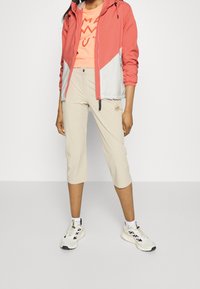 Chaqueta ligera rosa y gris con capucha; camiseta gráfica naranja; pantalones capri beige; zapatillas blancas con detalles negros.