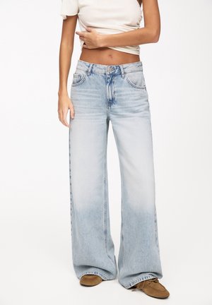 Femme portant un jean large bleu clair, un t-shirt blanc court et des chaussures marron, debout devant un fond blanc uni.