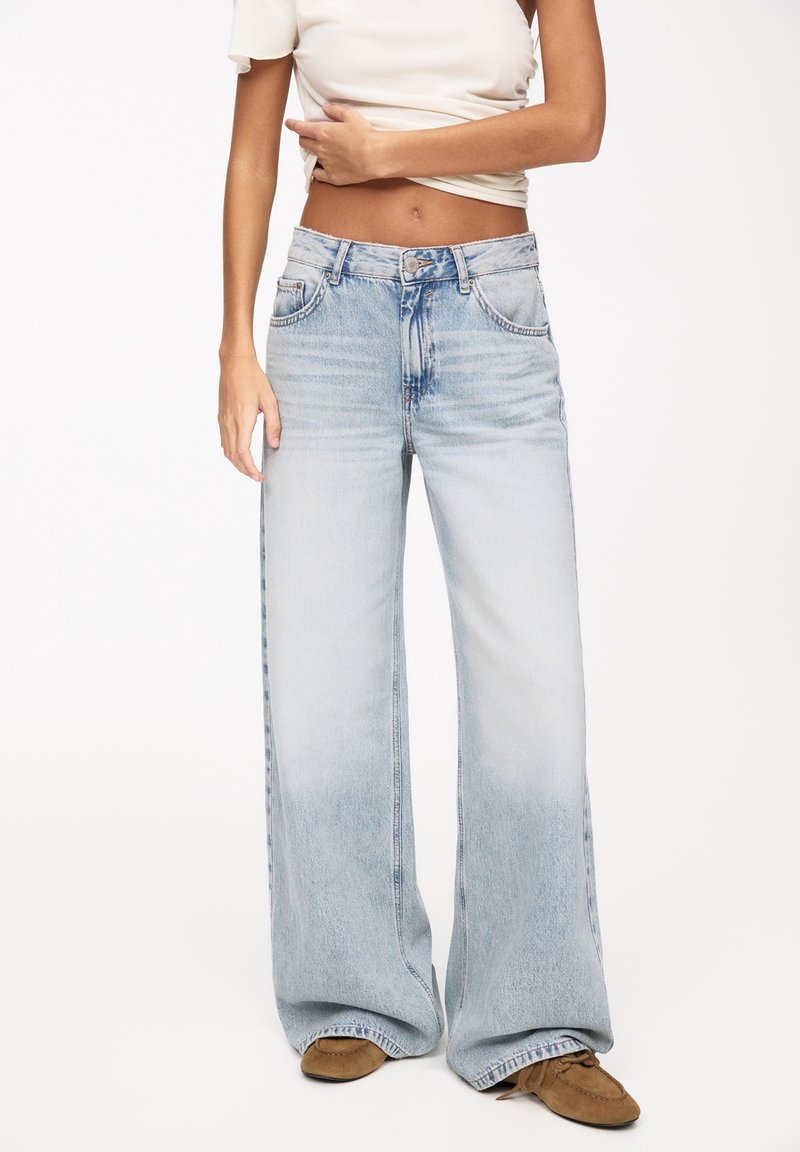 Femme portant un jean large bleu clair, un t-shirt blanc court et des chaussures marron, debout devant un fond blanc uni.