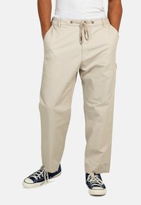 Beige, locker sitzende Hose mit elastischem Bund, Kordelzugverschluss und Seitentaschen, kombiniert mit dunkelblauen Sneakers.