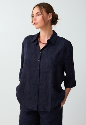 Vrouw met lichtbruin haar in een lage knot, draagt een donker gestructureerd overhemd met knopen en bijpassende broek, handen in de zakken, kijkt naar links.