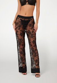 Juodos nėriniuotos kelnės su gėlių raštais, turinčios tvirtą juodą juosmenį, plačias kojas ir elegantišką juodą apdailą apačioje.