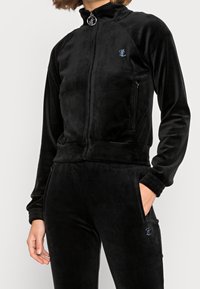 Black velour zip-up jacket with high collar, side zip pockets, and ribbed cuffs; features a small embroidered logo on the front.