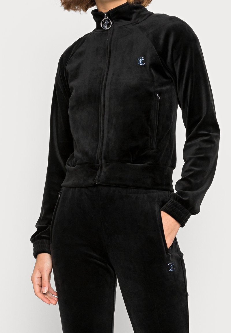 Black velour zip-up jacket with high collar, side zip pockets, and ribbed cuffs; features a small embroidered logo on the front.