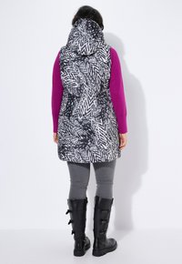 Gewatteerde vest met een zwart-wit zigzagpatroon, voorzien van een hoge kraag en capuchon, gedragen over een paarse lange mouw shirt en grijze leggings.