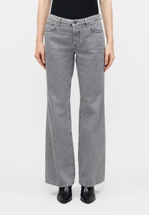 Žena oblečená v sivej džínsovej uniforme s vysokým pásom, širokými nohavicami a prednými vreckami, obutá v čiernych čižmách so špicatou špičkou, stojaca pred jednoduchým pozadím.