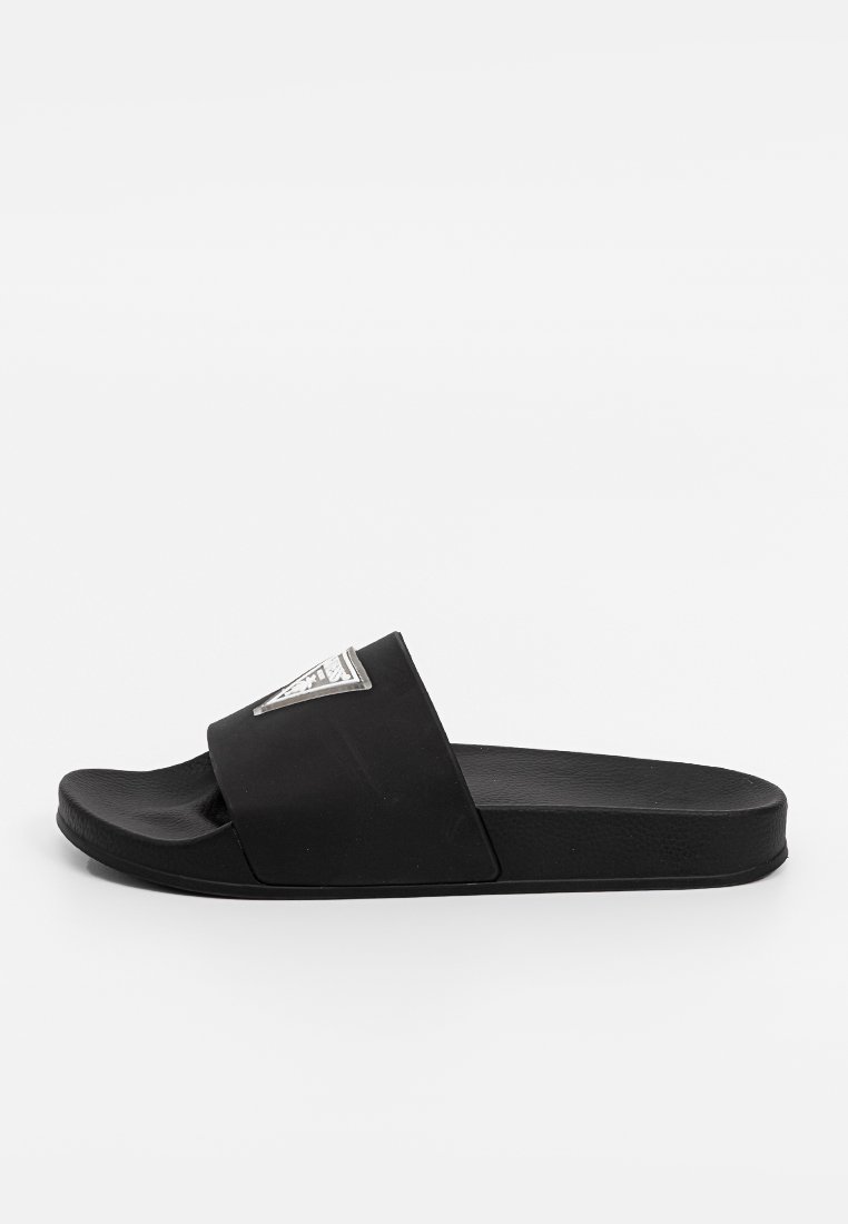 Sandalia deslizante negra de goma, con una amplia correa que presenta un acento de logo plateado en forma de triángulo. Textura suave, diseño de suela plana.