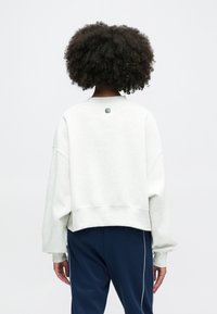 Svetlo šedý oversized mikinový sveter s voľne spustenými ramenami, prúžkovanými manžetami a lemom, s malou logó nášivkou na zadnej strane blízko goliera.