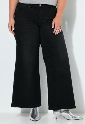 Person iført sorte vide jeans og sølvfarvede spidse ankelstøvler, der står mod en hvid baggrund.