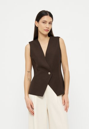 Γυναίκα με μακρύ σκούρο μαλλί, φορούν αμάνικο σκούρο καφέ γιλέκο με κουμπιά και άνετο λευκό παντελόνι, σε απλό φόντο.