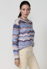 Pull en tricot multicolore avec un motif en vagues bleu, crème et rouille. Encolure ronde, manches longues et coupe ample. Texture légère.