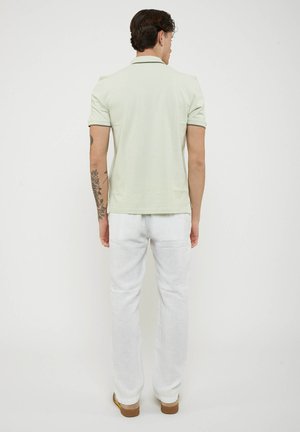 Uomo di spalle che indossa una polo verde chiaro, pantaloni bianchi, scarpe marroni e un tatuaggio floreale visibile sull'avambraccio sinistro.