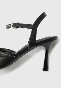 Zwarte geklede sandaal met een glanzende afwerking, slanke hak, verstelbare enkelband en metalen studs aan de voorkant.