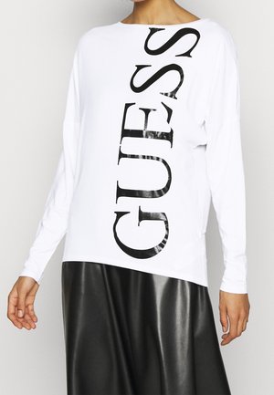 Persona con una camisa blanca de manga larga con un gran texto vertical negro que dice "GUESS" y una falda de cuero negra.
