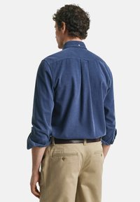 Dunkelblaues Button-Up-Hemd für Herren aus strukturiertem Stoff, mit einem Kentkragen, langen Ärmeln und einem Rückenfalt. Kombiniert mit beigen Hosen.