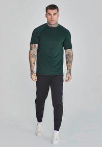 Zelená krátkopažná košeľa s textúrovanou látkou a raglanovými rukávmi, spárovaná s čiernymi joggers a bielymi teniskami.