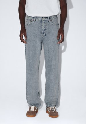 Mand iført lyseblå jeans med lige ben, hvid t-shirt og brune sneakers med hvide snørebånd, står mod en neutral baggrund.