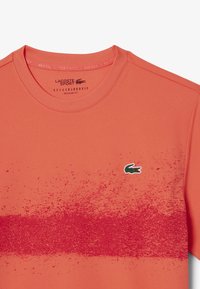 Kortärmad, korallfärgad träningsskjorta med gradientmönster i röd splatter. Har en grön Lacoste-logotyp och rundad halsringning. Tillverkad av mjukt tyg.