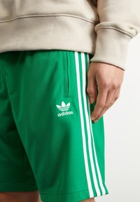 Gröna polyester sportshorts med vita vertikala ränder, med en Adidas-logotyp på vänster ben och en sidficka.