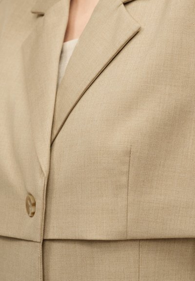 Μπεζ σακάκι φτιαγμένο στα μέτρα με ένα κουμπί και αιχμηλές πέτο, φορεμένο πάνω από ανοιχτόχρωμο τοπ.