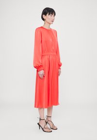 Korallfarbenes Kleid mit langen Ärmeln, tailliertem Schnitt, fließendem Rock und glatter Textur. Kombiniert mit schwarzen Riemchensandalen.