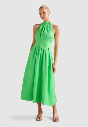 Žena v zelených plisovaných midi šatách bez rukávov s halterneck výstrihom a béžovými sandálmi s otvorenou špičkou stojí na jednoduchom pozadí.