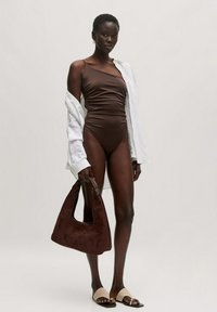 Bruin één stuk badpak met een enkele schouderband, gecombineerd met een losse witte blouse. Bruin suède handtas en beige instappers.