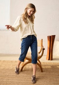 Frau lächelt und balanciert auf einem Fuß, trägt eine blumige Bluse, knielange Jeansshorts und braune Sneakers in einem schlichten Raum mit einem hölzernen Stuhl.