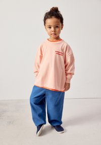Jeune enfant aux cheveux attachés porte un sweat-shirt pêche surdimensionné, un pantalon bleu et des chaussures sombres sans lacets, debout devant un fond uni.
