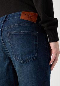 Tamno plave traperice s teksturiranom završnom obradom. Posjeduju smeđu kožnu oznaku Calvin Klein na pojasu i standardne stražnje džepove.