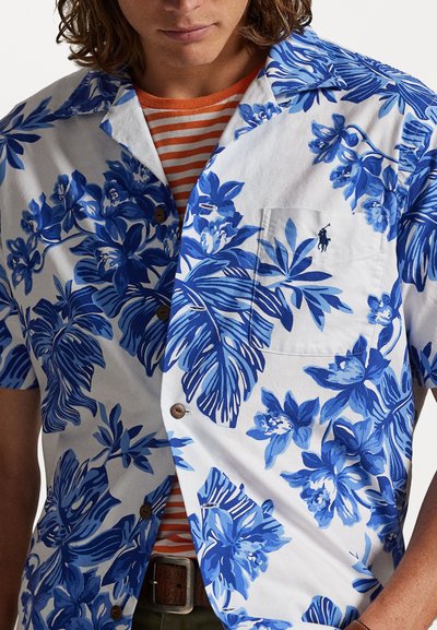 Κοντομάνικο, λευκό πουκάμισο με μπλε λουλουδάτο μοτίβο και καφέ κουμπιά. Φορεμένο πάνω από ένα πορτοκαλί και λευκό ριγέ πουκάμισο, το οποίο διαθέτει τσέπη στο στήθος.