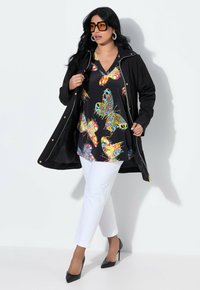 Svart lättviktsjacka över en flerfärgad tops med fjärilsmönster, i kombination med vita byxor och svarta klackar, som framhäver livfulla mönster.