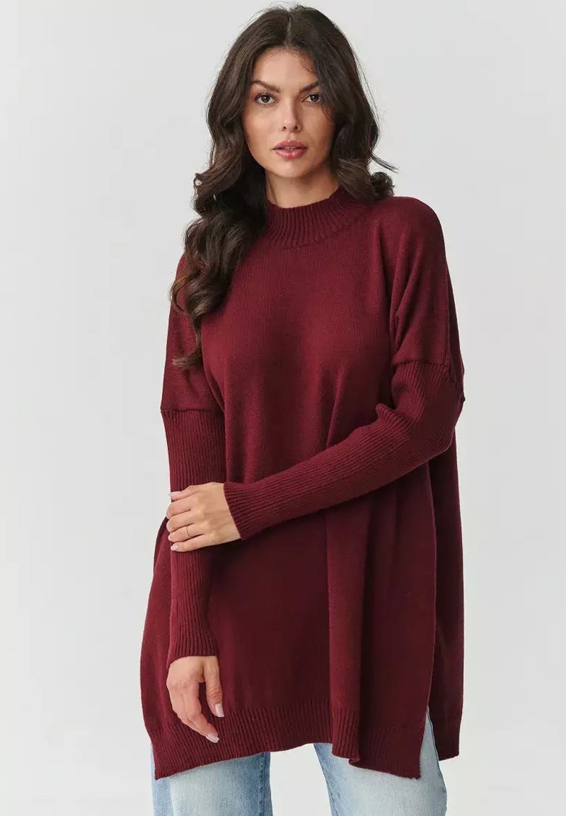Burgundowy, oversize sweter o prążkowanej fakturze, z wysokim kołnierzem i długimi rękawami, połączony z jasnoniebieskimi dżinsami.