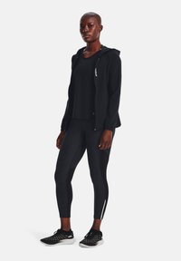 Chaqueta atlética negra con capucha, camiseta negra ajustada, leggings negros texturizados y zapatillas de running negras con acentos en blanco y detalles de malla.