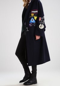 Abrigo de lana azul marino con grandes parches bordados, diseño abierto, mangas largas y un corte relajado; combinado con pantalones ajustados negros y botines.