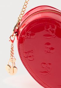 Κόκκινη τσάντα χειρός από δερμάτινο λάκα σε σχήμα καρδιάς με ανάγλυφο σχέδιο κερασιού, χρυσά μεταλλικά στοιχεία, αλυσιδάκι και κρεμαστό διακοσμητικό σε σχήμα καρδιάς.