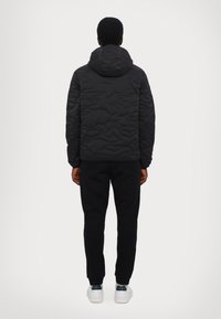 Sort quiltet jakke med hætte, der har en struktureret overflade. Båret over sorte bukser og hvide sneakers. Bagfra vises et enkelt design.